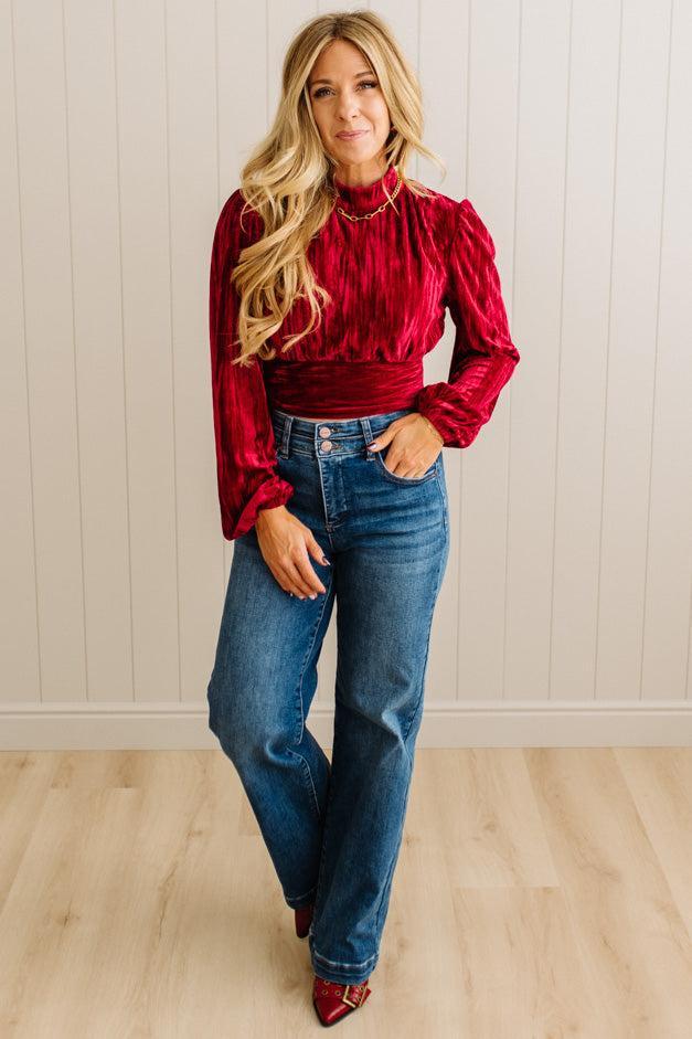 Woman wearing a red blouse and blue jeans standing against a white paneled wall.