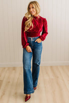 Woman wearing a red blouse and blue jeans standing against a light wooden paneled wall.