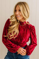 Woman wearing a red velvet blouse with long sleeves and blue jeans against a neutral background