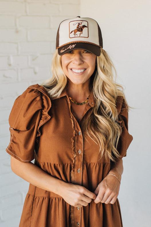 Woman wearing a brown dress and cap with a horse design against a white brick wall.