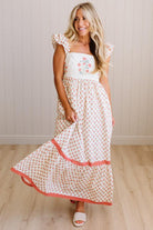 Woman wearing a white dress with floral embroidery and red accents, standing against a light wooden paneled wall.