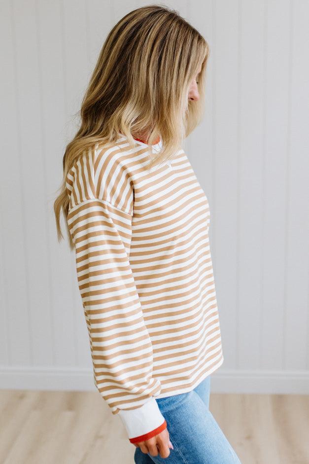 BLONDE WOMAN WEARING A CAMEL AND WHITE STRIPED CREW NECK PULLOVER WITH WHITE AND DARK ORANGE KNIT RIB BAND TRIMMED, IT IS LONG SLEEVED WITH A RELAXED FIT. SHE IS STANDING ON A LIGHT WOOD FLOOR WITH LIGHT BACKGROUND.

