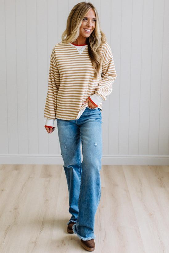 BLONDE WOMAN WEARING A CAMEL AND WHITE STRIPED CREW NECK PULLOVER WITH WHITE AND DARK ORANGE KNIT RIB BAND TRIMMED, IT IS LONG SLEEVED WITH A RELAXED FIT. SHE IS STANDING ON A LIGHT WOOD FLOOR WITH LIGHT BACKGROUND.
