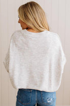 A woman viewed from behind wearing a light heather gray oversized crewneck sweater and blue jeans, standing against a light beige paneled wall.