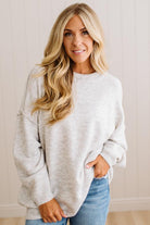 A woman with long blonde hair wearing an oversized light heather gray crewneck sweatshirt and blue jeans, standing against a light beige paneled wall.