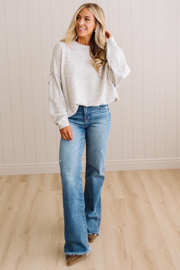 A woman modeling a light heather gray oversized knit sweater and blue flare jeans with a frayed hem, standing on a light wood floor against a beige paneled wall.