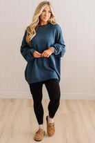 A woman modeling an oversized blue knit sweater with black leggings and tan slip-on shoes, standing against a white paneled wall and light wood floor.