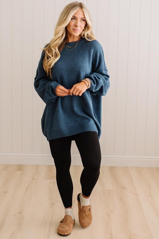 A woman modeling an oversized blue knit sweater with black leggings and tan slip-on shoes, standing against a white paneled wall and light wood floor.