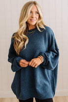 A woman with long blonde hair wearing an oversized, dusty slate-blue knit sweater, standing against a white paneled wall.