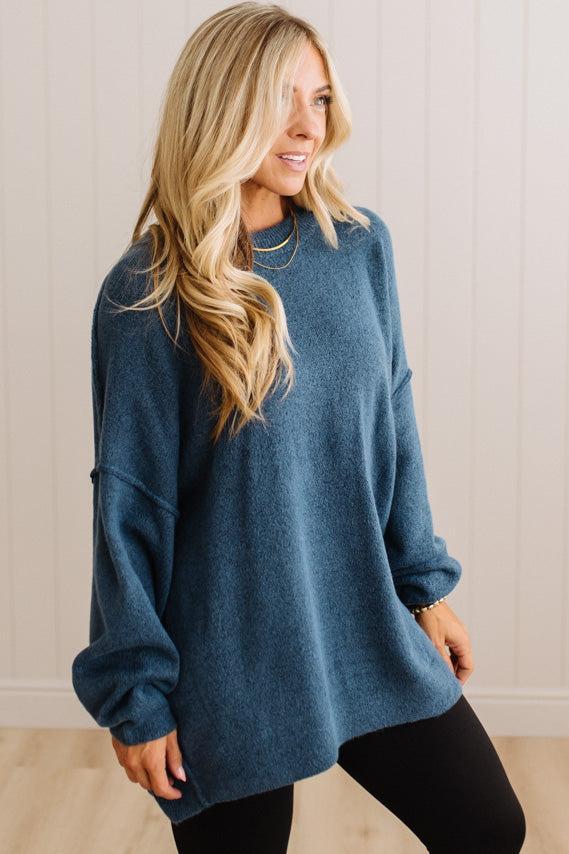 A woman wearing a blue knit crewneck sweater and black leggings, standing against a light beige paneled wall.
