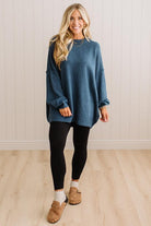 A woman modeling an oversized blue knit sweater with black leggings and tan slip-on shoes, standing against a white paneled wall and light wood floor.