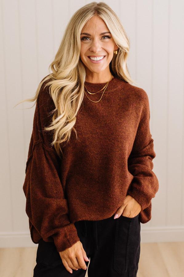 A smiling blonde woman wearing a fuzzy, oversized brown crewneck sweater and black pants, standing against a light beige paneled wall.