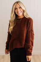 A smiling blonde woman wearing a fuzzy brown long-sleeve sweater and black pants, standing against a light beige paneled wall.