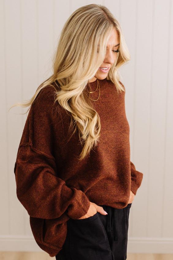 A woman wearing a brown knit crewneck sweater and black pants, standing against a white paneled wall.
