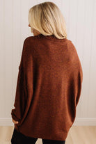 A woman with blonde hair is shown from behind wearing a loose-fitting, rust-brown knit sweater and black pants, standing against a white paneled wall.