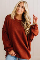 A smiling blonde woman wearing a rust oversized crewneck sweatshirt and blue jeans, standing against a light beige paneled wall.