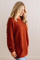 A woman wearing a fuzzy, oversized rust-orange crewneck sweater, standing against a light beige paneled wall.