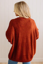 A woman viewed from behind wearing a loose, oversized rust-orange knit sweater and blue jeans, standing against a white paneled wall.