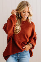 A woman wearing a cozy, oversized rust-orange knit sweater and light-wash blue jeans, standing against a light beige paneled wall.