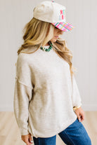 A woman with long blonde hair wearing an oversized light heather biege crewneck sweatshirt and blue jeans, standing against a light beige paneled wall.