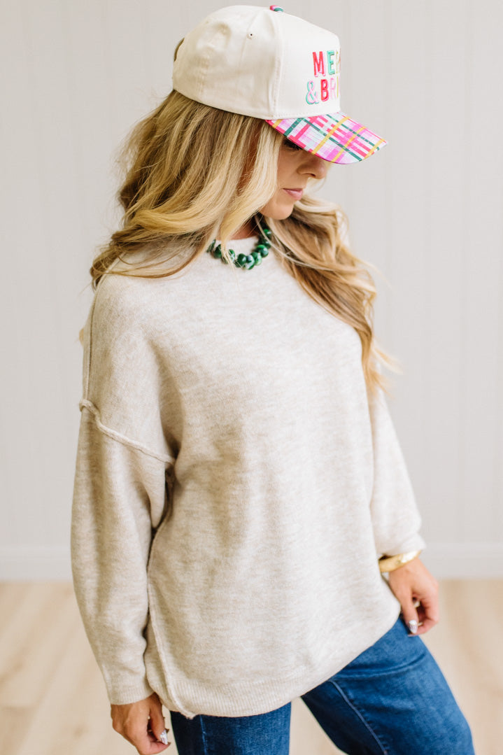 A woman with long blonde hair wearing an oversized light heather biege crewneck sweatshirt and blue jeans, standing against a light beige paneled wall.