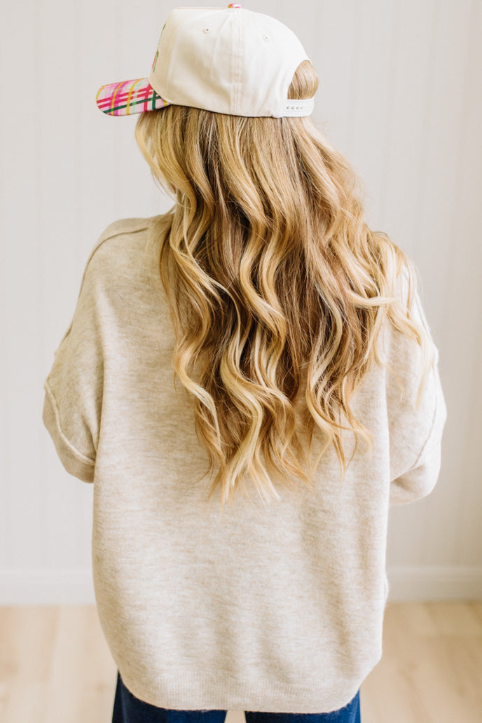 A woman with long blonde hair wearing an oversized light heather beige crewneck sweatshirt and blue jeans, standing against a light beige paneled wall.