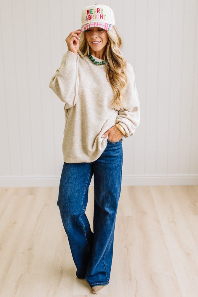 A woman with long blonde hair wearing an oversized light heather beige crewneck sweatshirt and blue jeans, standing against a light beige paneled wall.