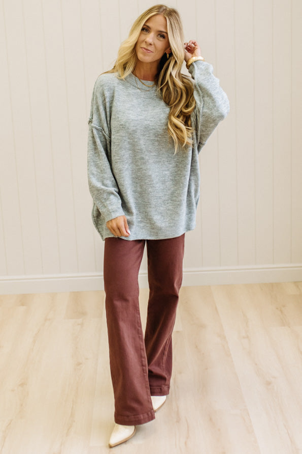 A woman with long blonde hair wearing an oversized light heather gray crewneck sweatshirt and blue jeans, standing against a light beige paneled wall.
