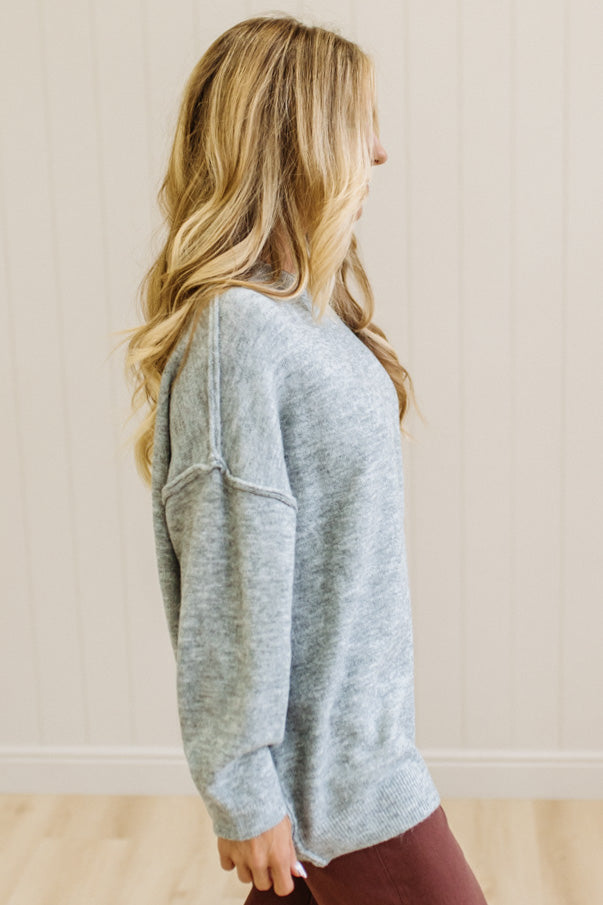 A woman with long blonde hair wearing an oversized light heather gray crewneck sweatshirt and blue jeans, standing against a light beige paneled wall.