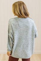 A woman with long blonde hair wearing an oversized light heather gray crewneck sweatshirt and blue jeans, standing against a light beige paneled wall.