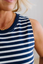 Blonde woman wearing a navy blue and white horizontal striped tank top knee length dress with sandals, standing on a wood floor with neutural backdrop.
