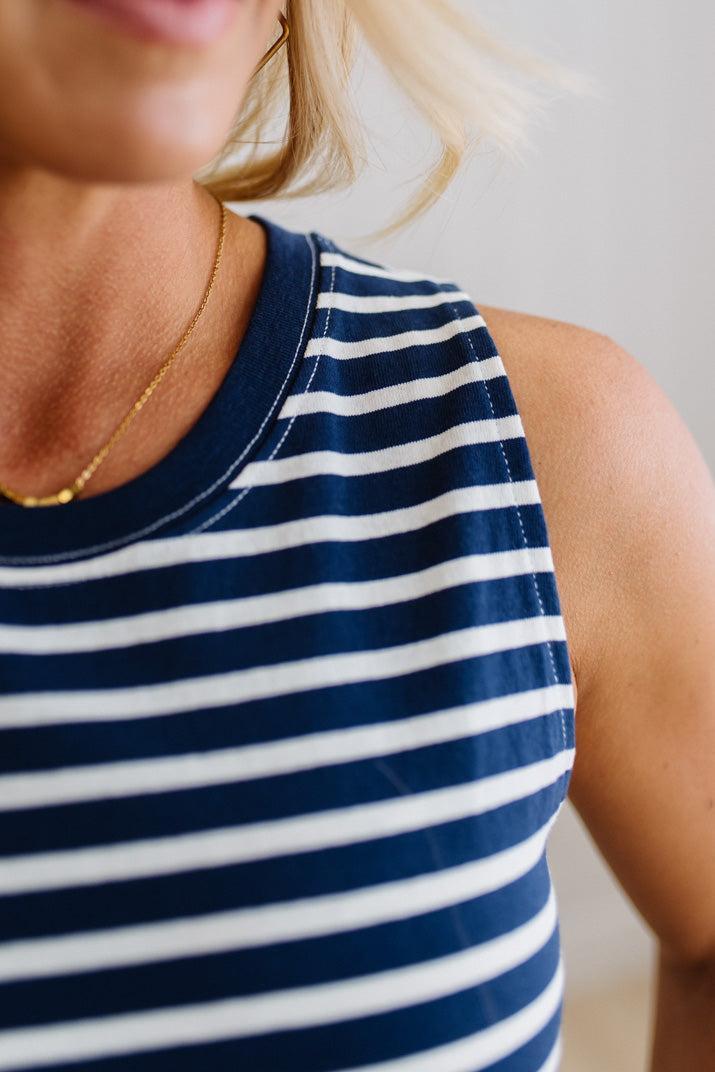 Blonde woman wearing a navy blue and white horizontal striped tank top knee length dress with sandals, standing on a wood floor with neutural backdrop.