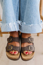 Brown sandals with buckles worn with light blue jeans on a neutral background