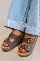 Brown sandals with buckles worn with rolled-up blue jeans on a neutral background
