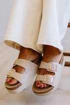 Beige platform slide sandals with double buckled straps and gold-tone hardware, shown on a person's feet wearing cream-colored pants.