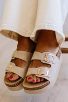 Beige double-strap platform slide sandals with gold-tone buckle details, shown on a model wearing white pants.