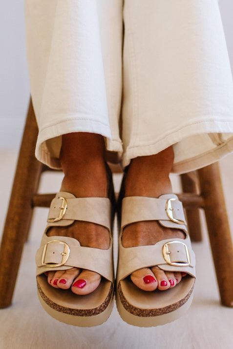 Beige double-strap platform slide sandals with gold-tone buckles, shown on a model's feet wearing cream-colored pants.