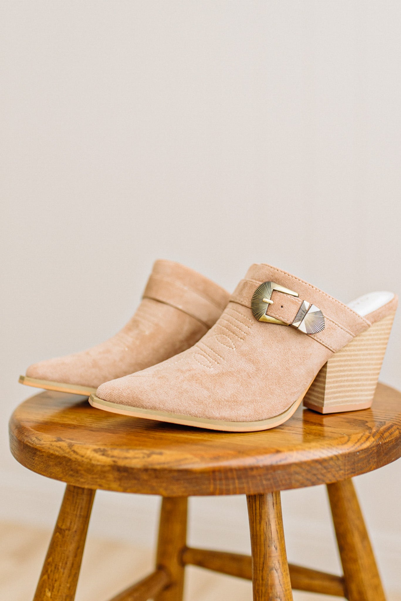 Pair of beige suede mules with a wooden stool on a neutral background