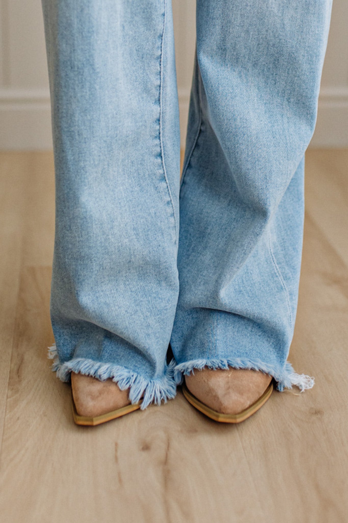 Light blue jeans with frayed cuffs worn by a person on a wooden floor.