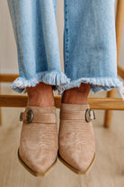 Close-up of beige suede shoes with metallic buckles worn with light blue jeans.