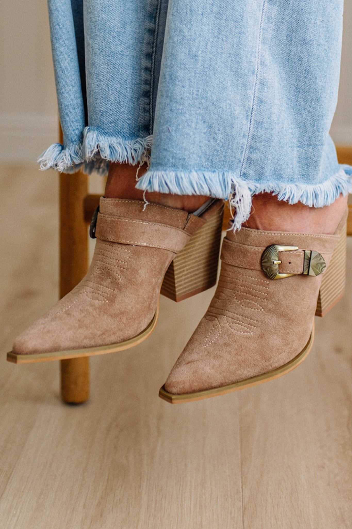 Tan suede ankle boots with gold buckles worn with light blue jeans on a wooden floor.