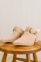 Beige suede ankle boots with gold buckles on a wooden stool against a beige background
