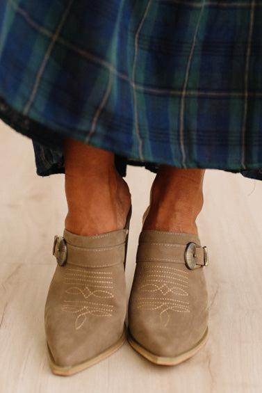 A close-up of a person wearing taupe suede mule boots with western-style stitching and buckle straps, paired with a blue plaid skirt on a light wood floor.