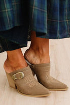 A close-up of a person wearing taupe suede pointed-toe mule boots with a stacked block heel, decorative stitching, and a gold buckle detail, paired with a blue plaid skirt on a light wood floor.