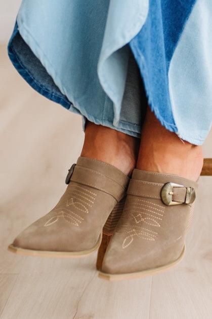 A close-up of a person's feet wearing taupe suede mule boots with decorative stitching and buckle straps, paired with denim two tone jeans on a light wood floor.