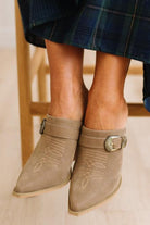 A close-up of a person's feet wearing taupe suede mule boots with decorative stitching and buckle straps, paired with a blue plaid skirt.