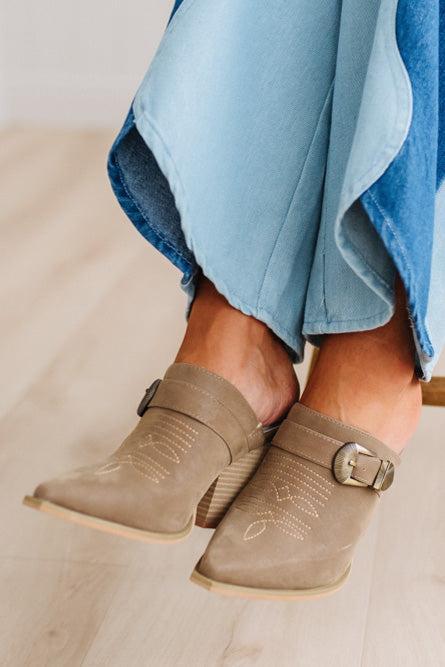 A close-up of a person's feet wearing taupe suede mule boots with decorative stitching and buckle straps, paired with denim two tone jeans on a light wood floor.