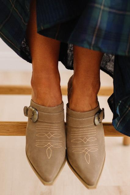 A close-up of a person's feet wearing taupe pointed-toe ankle boots with decorative stitching and buckle straps, paired with a blue plaid skirt.