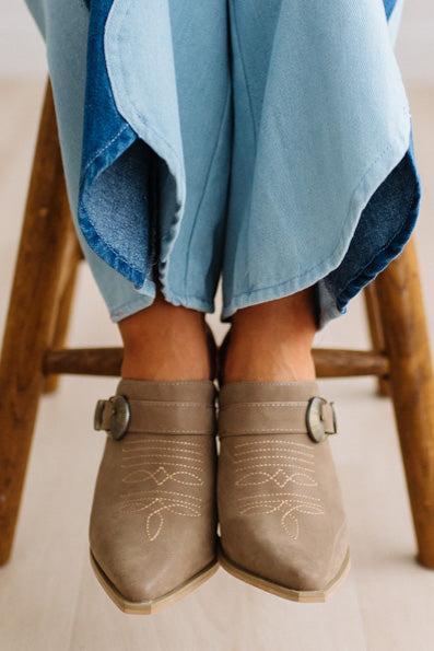 A close-up of a person's feet wearing taupe suede mule boots with decorative stitching and buckle straps, paired with denim two tone jeans on a light wood floor.