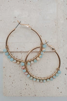 Gold hoop earrings with small light blue, pink, and white beads along the lower edge, displayed on a light beige stone surface.